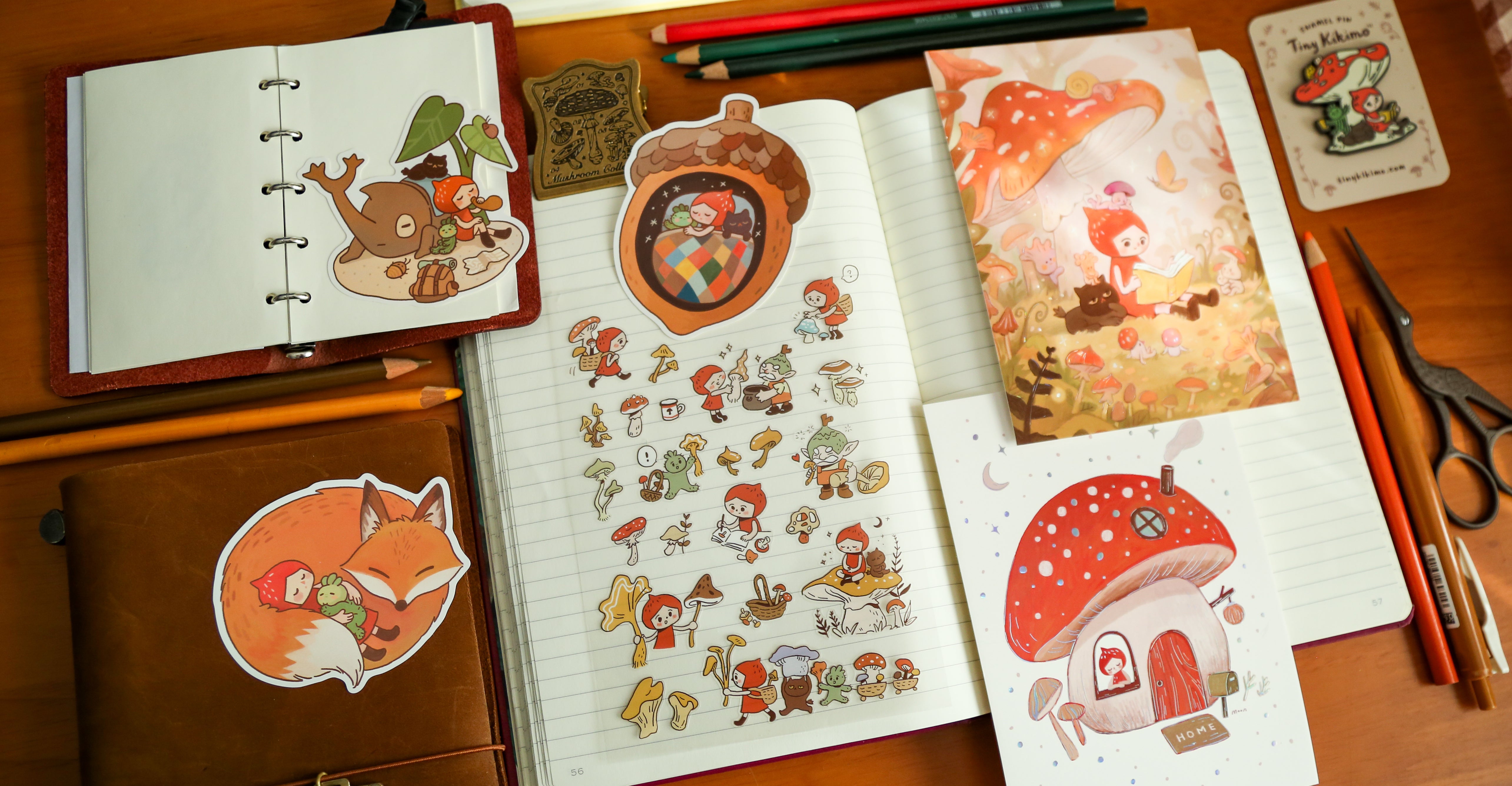 A notebook with Tiny Kikimo stickers on a wooden surface with stationery items.