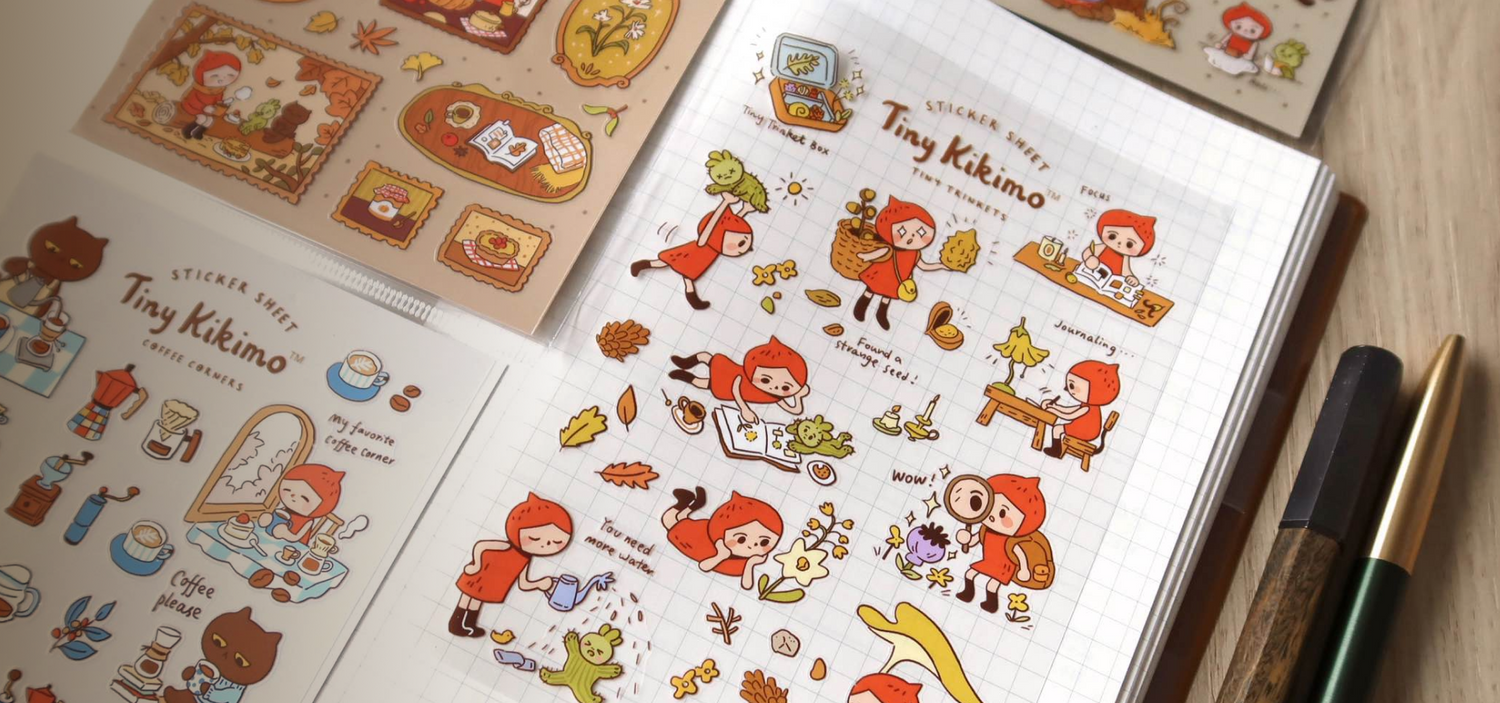 Sticker sheets with Tiny Kikimo on a wooden surface with a pen and pencil.